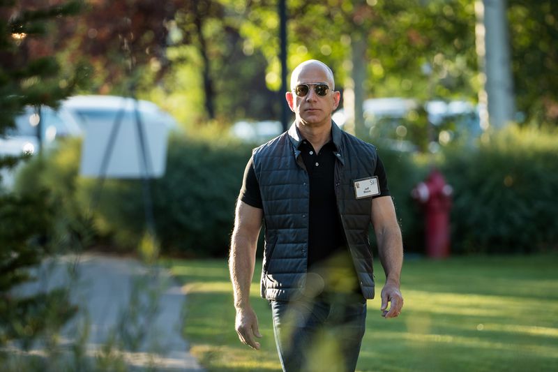 Jeff Bezos wears aviator sunglasses, a quilted black vest with a laminated name tag, a fitted, black polo shirt, and black, slim-fitting pants. His arms are noticeably muscular. 