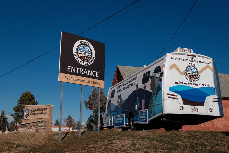 Several exterior signs for Oyate Health Center next to a mobile healthcare unit bearing the health center’s logo under a blue sky.