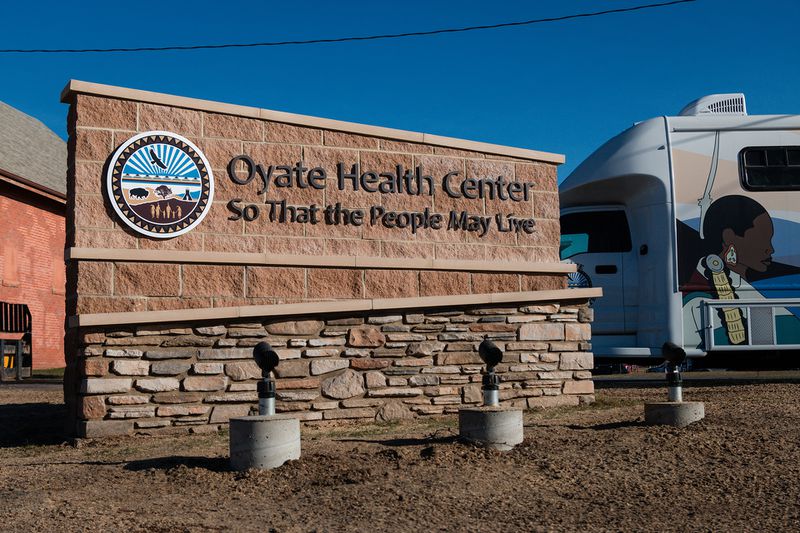 An exterior rock-and-brick sign reading “Oyate Health Center” and “So That The People May Live” alongside the Great Plains Tribal Leaders’ Health Board logo against a blue sky.