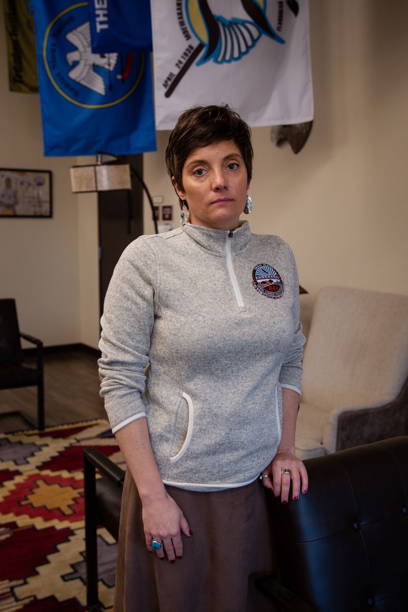 A woman with short hair wearing a sweater with the Great Plains Tribal Leaders’ Health Board logo stands indoors under tribal flags.