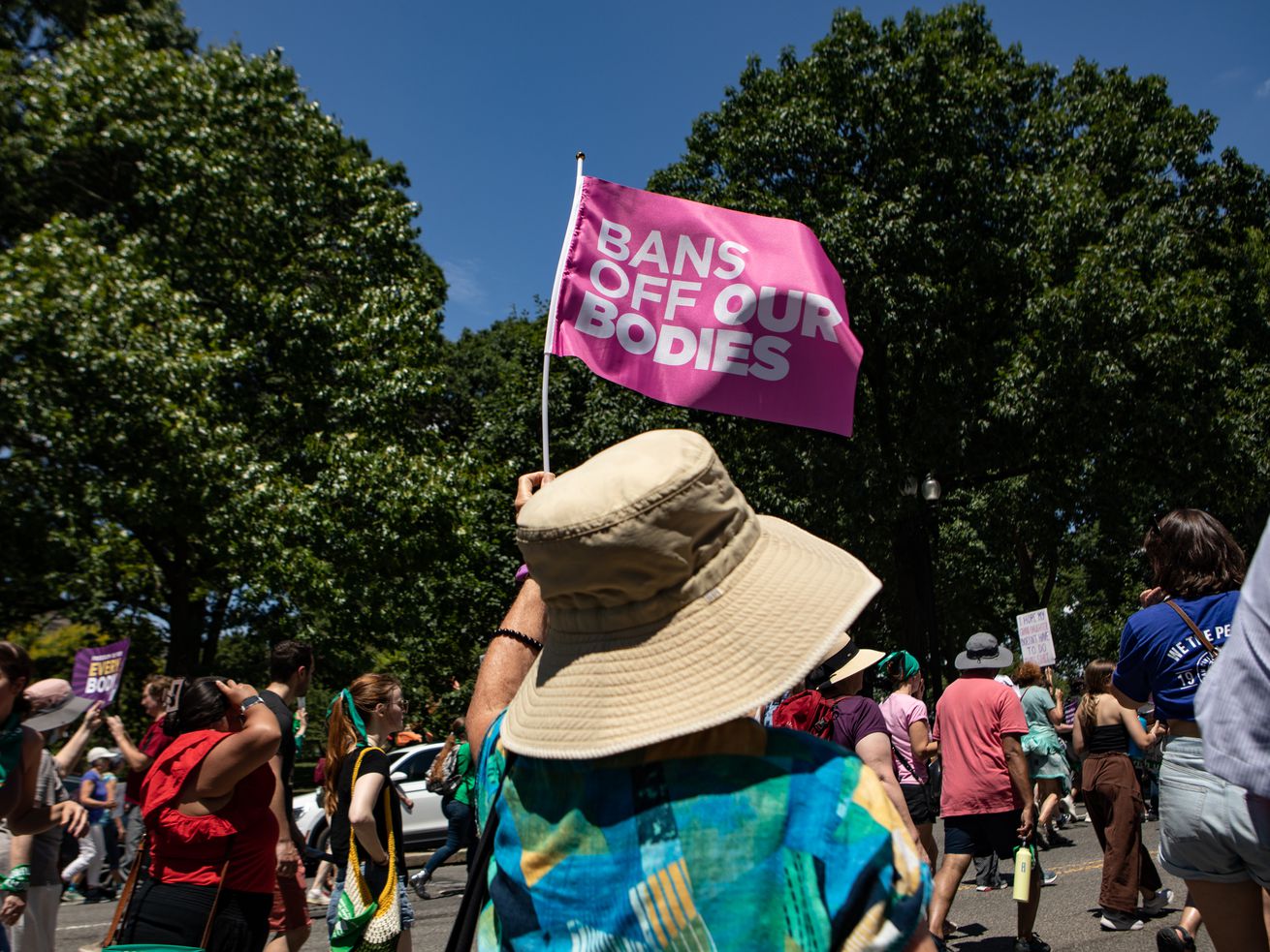 A person in a sun hat walks in a crowd waving a flag that reads “Bans off our bodies.”