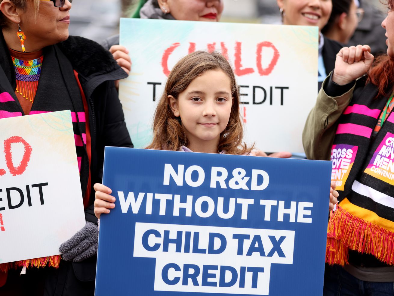 A child in a crowd holding up a sign that says NO R&amp;D WITHOUT THE CHILD TAX CREDIT.