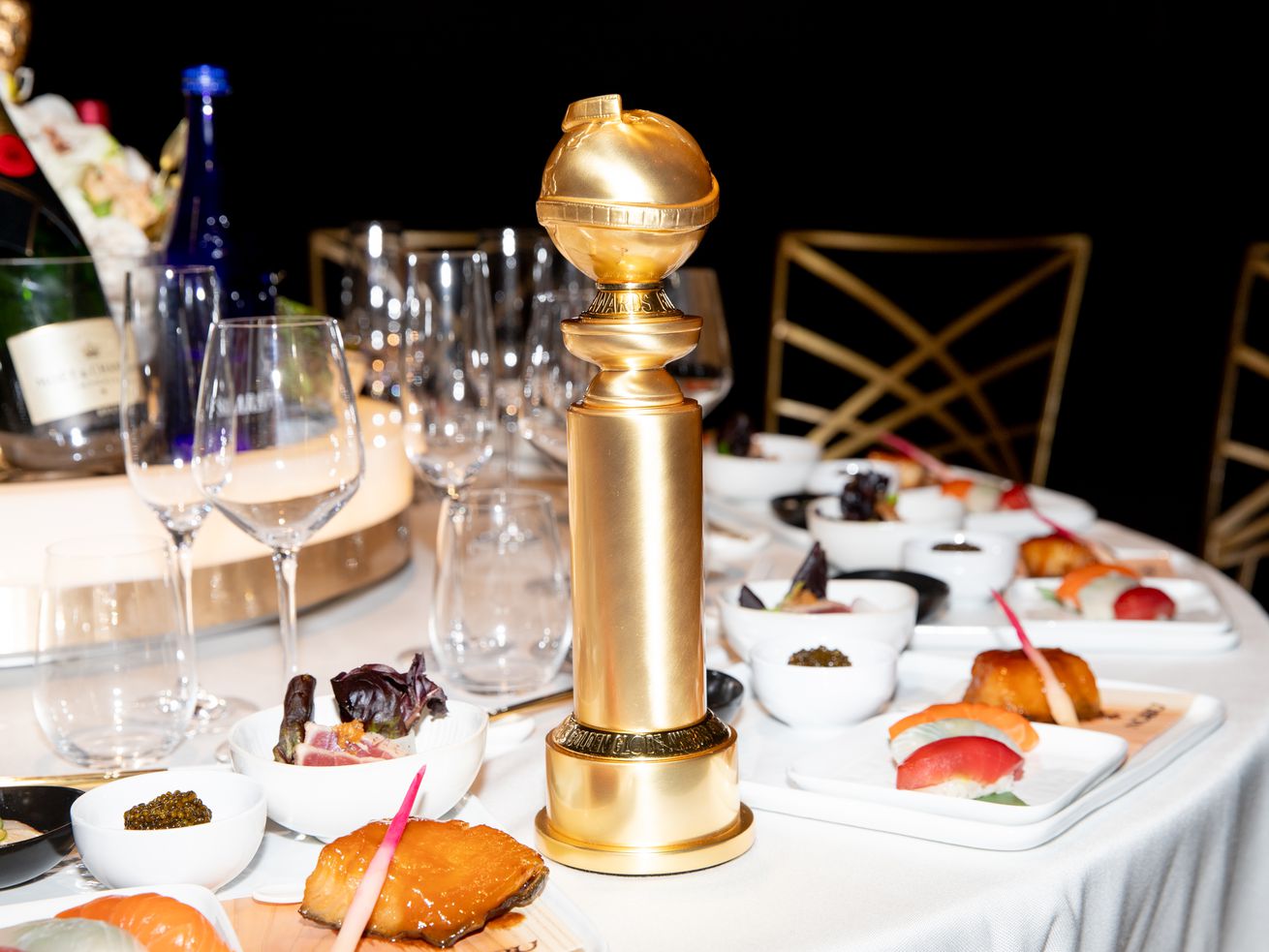 A Golden Globe statue standing on a dining table with a white tablecloth, dishes of food, and a bottle of champagne.