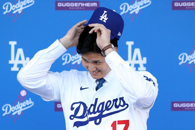 Ohtani wears a white Dodger’s uniform and smiles as he puts on a blue LA dodgers baseball cap.