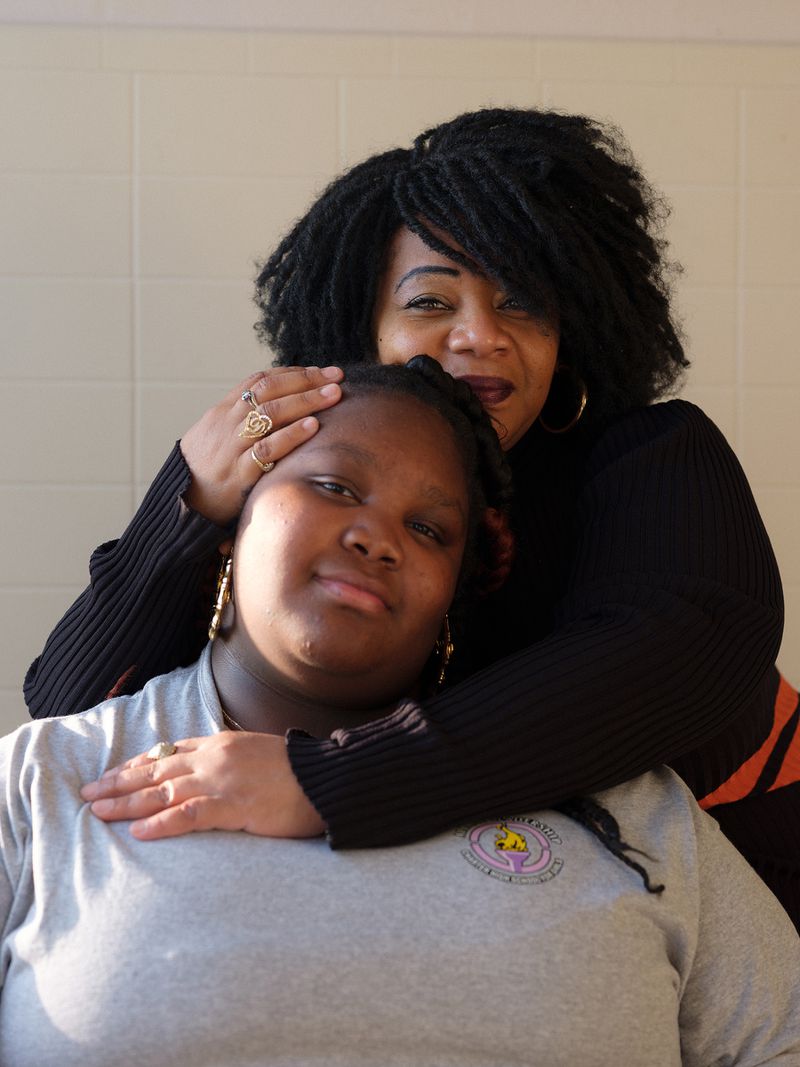 A Black woman and teenage girl pose for a portrait, the girl sitting while the woman hugs her around the shoulders from behind. Both are smiling at the camera.