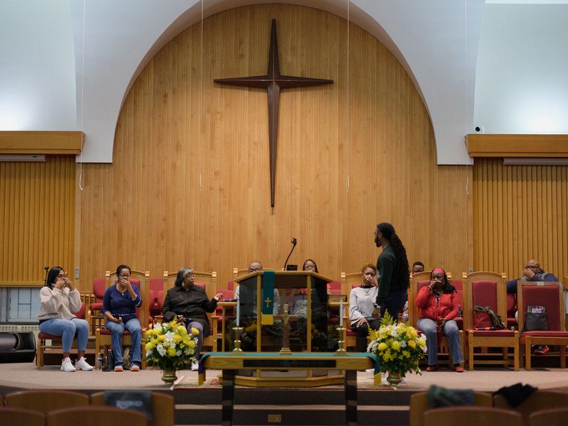 People sit in rows of red chairs at the front of a church sanctuary, with a Black man standing in front of them leading them in song.