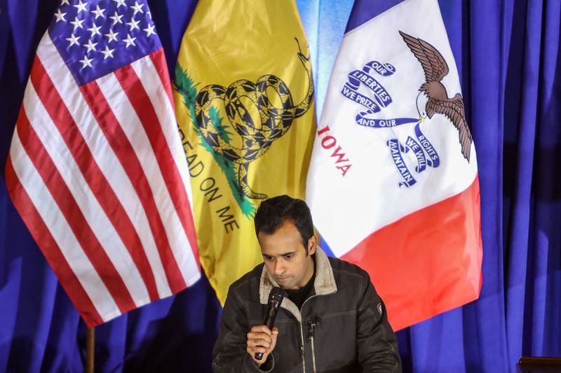 Vivek Ramaswamy holding a microphone in front of a backdrop of an American flag, a Don’t Tread On Me flag, and an Iowa flag.