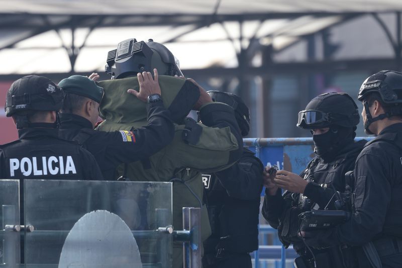 People in heavy tactical gear help their colleague into a bomb-proof suit. One person has the word “Policia” visible on the back of their jacket.