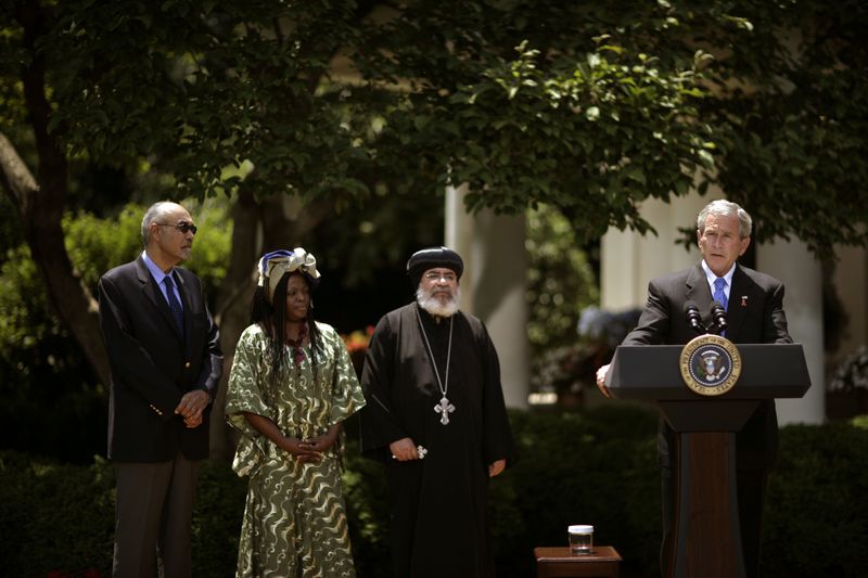 President Bush speaks at a microphone while Jean William “Bill” Pape, “Aunt” Manyongo Mosima “Kuene” Tantoh, and Bishop Paul Yowakim look on.