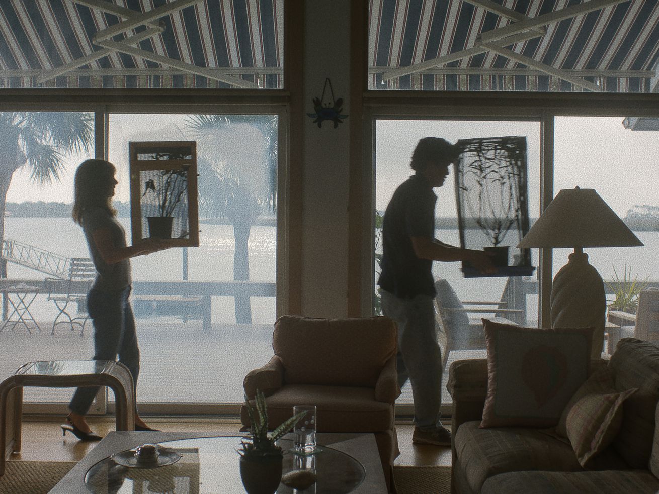 A scene from the movie May December shows a woman and a man, separated by several feet, walking through a home in front of large glass windows, each carrying a plant.