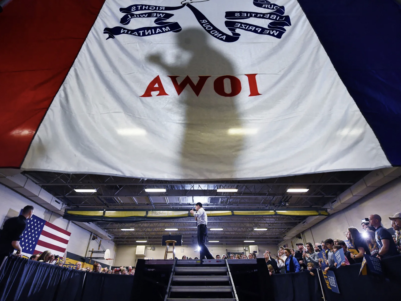 A shadow of a man is cast on a large white banner reading “Iowa” hanging in a school gym, where the middle of the room is a stage platform and the man is walking onto it.