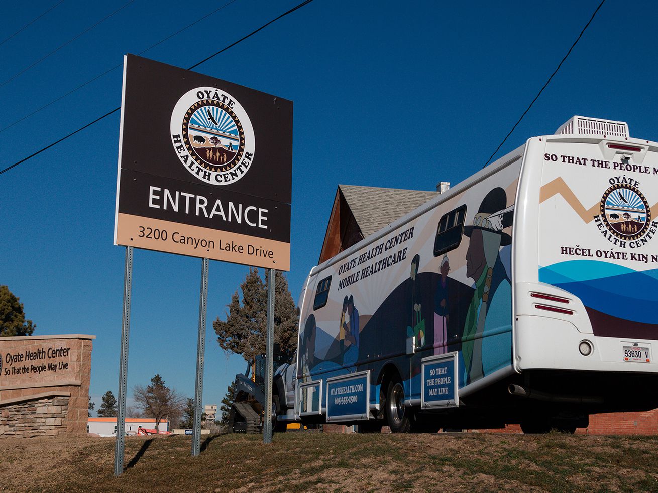 Several exterior signs for Oyate Health Center next to a mobile health care unit bearing the health center’s logo under a blue sky.