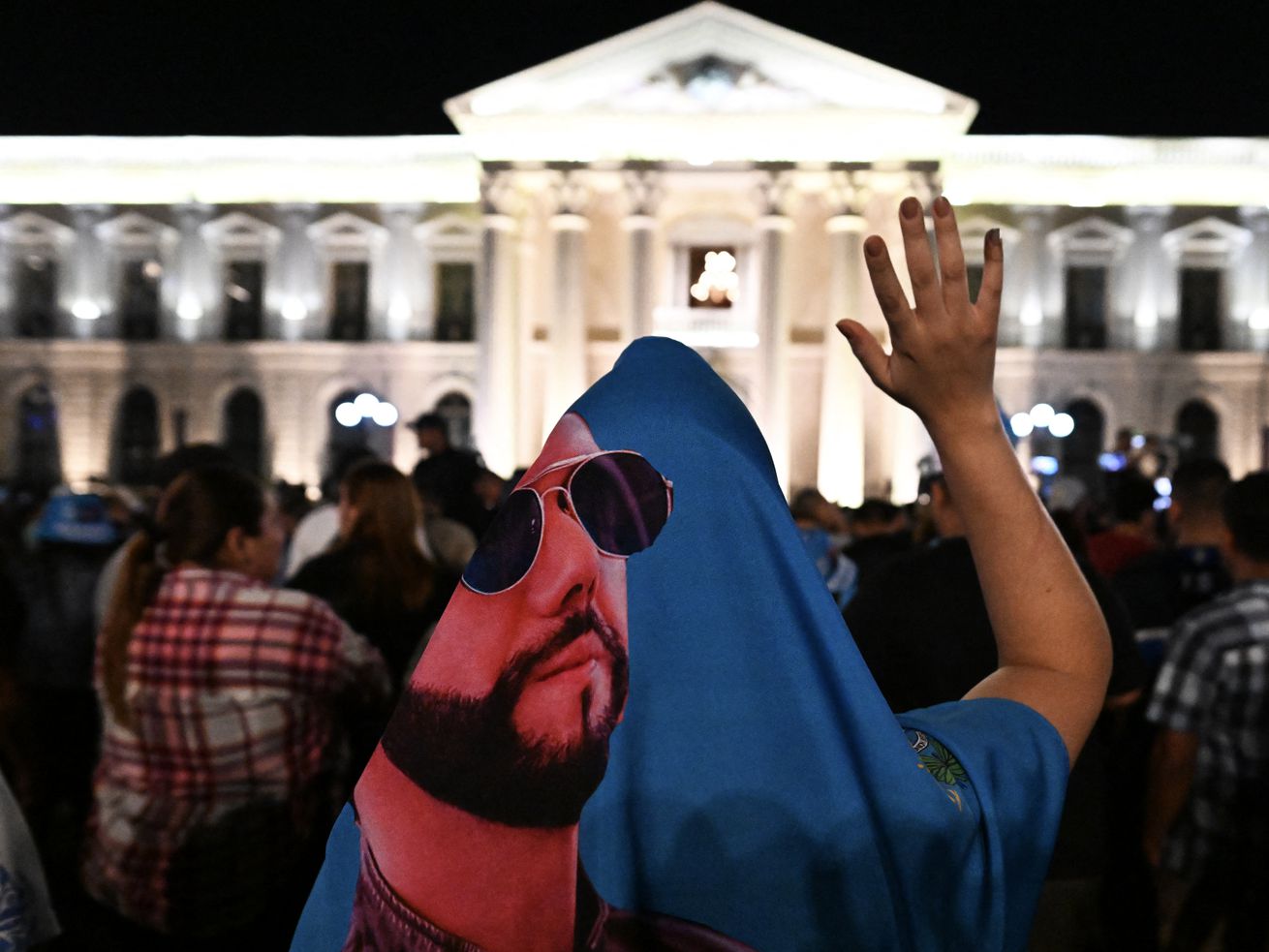 Bukele supporters gather outside the National Palace in San Salvador at night. The person closest to the camera is draped in a printed image of Bukele wearing sunglasses.