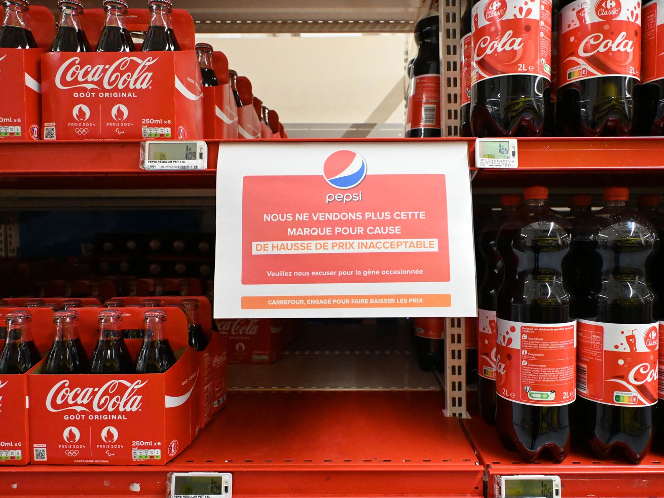 Grocery shelves with Coca-Cola bottles, with an empty space where Pepsi’s products would typically go. A sign written in French informs customers that some PepsiCo products are not available due to unacceptable price increases.